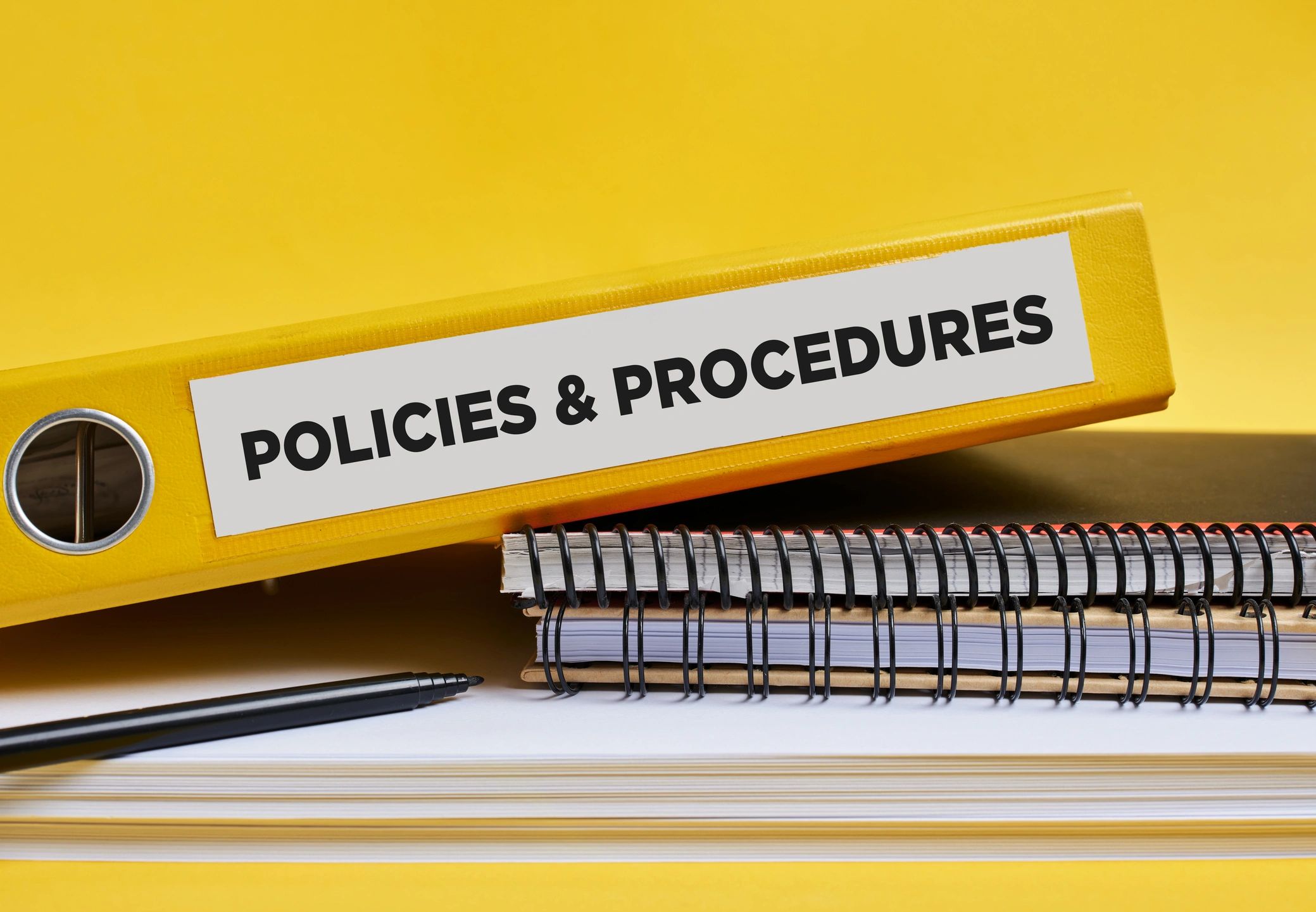 Spiralli defterlerin ve kâğıtların üzerinde duran, “Policies & Procedures” etiketli sarı klasör ve bir keçeli kalem